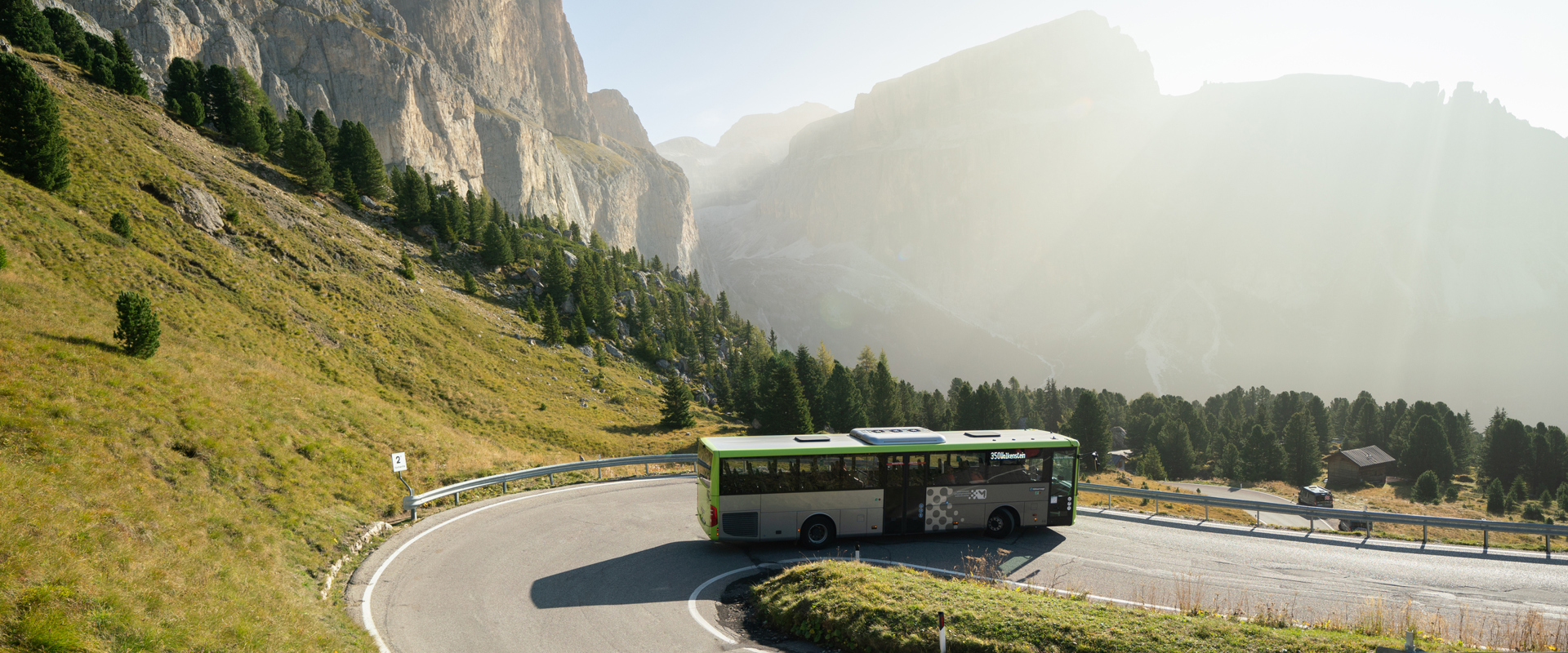 By bus through the Dolomites Bus driving downhill on a bend, a sunny mountain landscape with meadows, trees and rock faces in the background